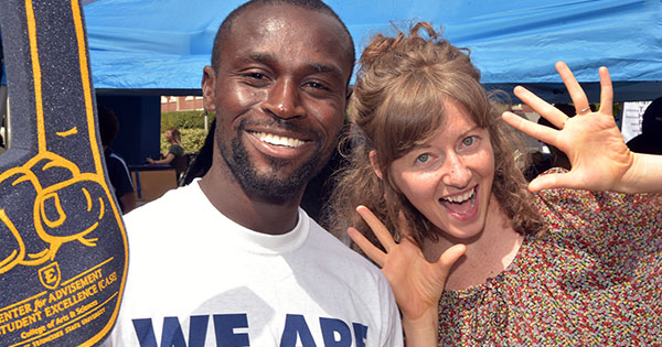 A young man and woman pose for the camera. The man is holding a foam finger and the woman is giving jazz hands.