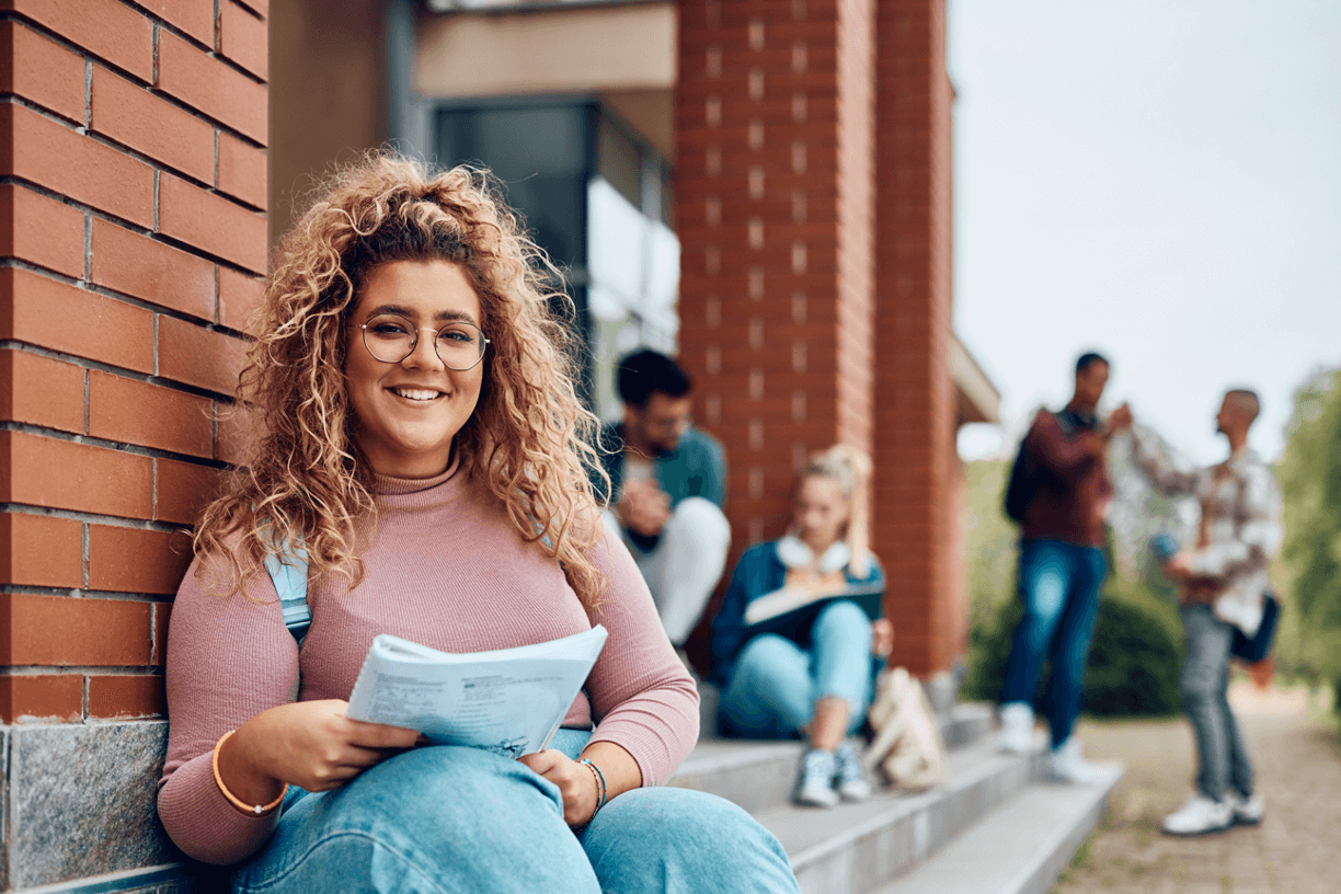 A young woman sits on steps outside a brick building on a college campus. 