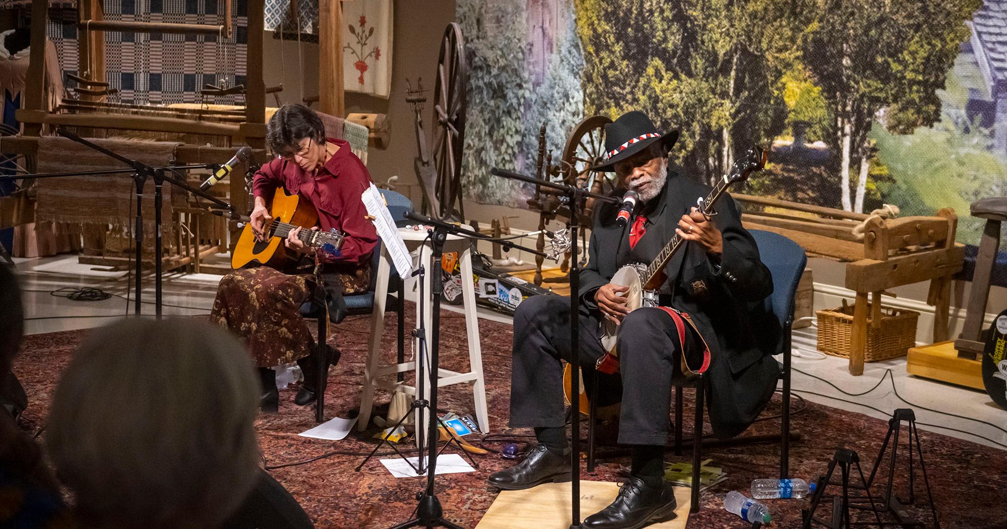 Image depicts two people playing guitar in front of an audience.