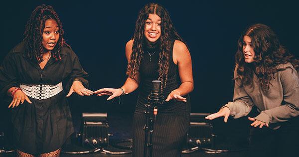 3 singers performing on stage and sharing a microphone