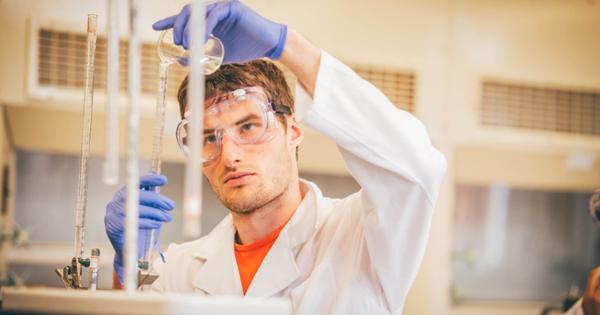 A chemistry student wearing protective goggles and gloves pours liquid into a beaker.