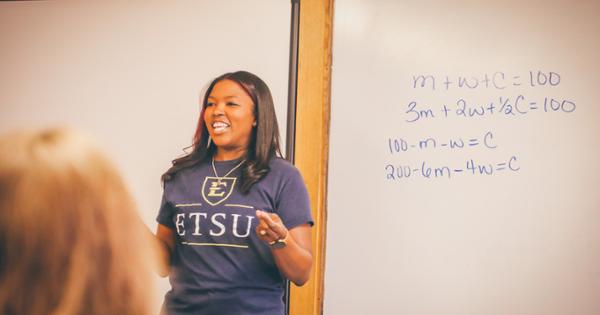 A teacher standing at the front of the classroom in front of a white board. On the board are mathematical equations.