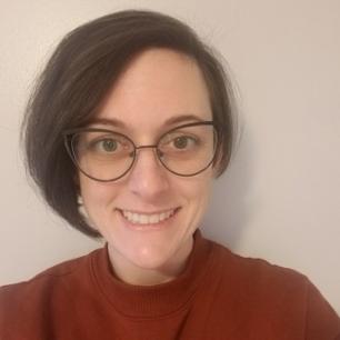 A white woman with chin-length, straight brown hair, horn-rimmed glasses, and a terracotta colored sweater smiles into the camera. of Rebecca Strasser