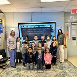 Two female teachers and a group of students smiling in front of a classroom board containing the class mission statement: "I am a reader and a thinker and a leader! I will work hard and help others. We are a classroom family."