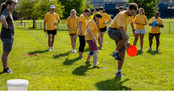 A group of students in matching yellow shirts participate in a team-building activity on a grassy field. In the foreground, one student balances while reaching toward a bright orange disc as classmates watch and wait their turn. A facilitator stands nearby observing the activity.
