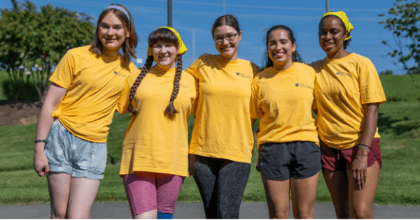 Five students wearing matching yellow T-shirts stand arm in arm outdoors, smiling at the camera. The group poses on a paved path with a grassy hillside and trees in the background on a sunny day.