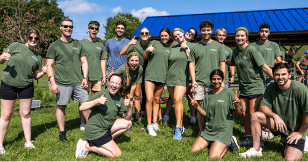A group of students wearing matching green T-shirts pose together outdoors, smiling and giving thumbs-up and peace signs toward the camera. The group stands on a grassy field near a blue-roofed pavilion during a sunny day.
