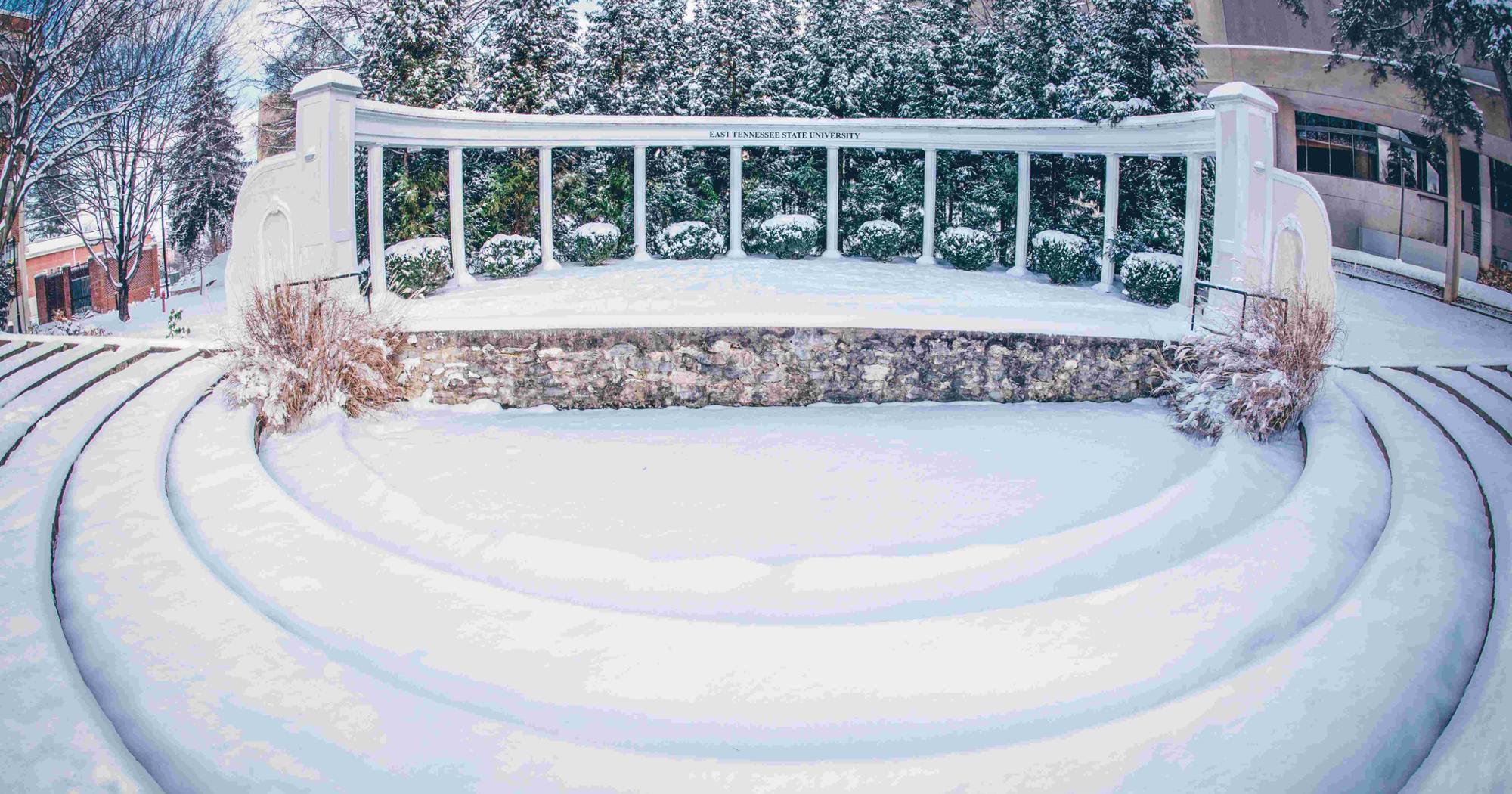 A snow-covered amphitheater at East Tennessee State University with a curved stone stage and white columns set against evergreen trees blanketed in snow.