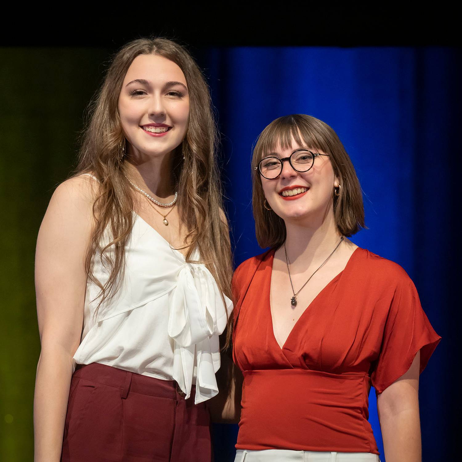 Two women (one wearing red pants and a white blouse and another in white pants and a red blouse) smile into the camera while side hugging.