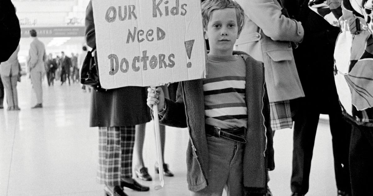 Old photo of a child holding a sign that reads “Our kids need doctors!”