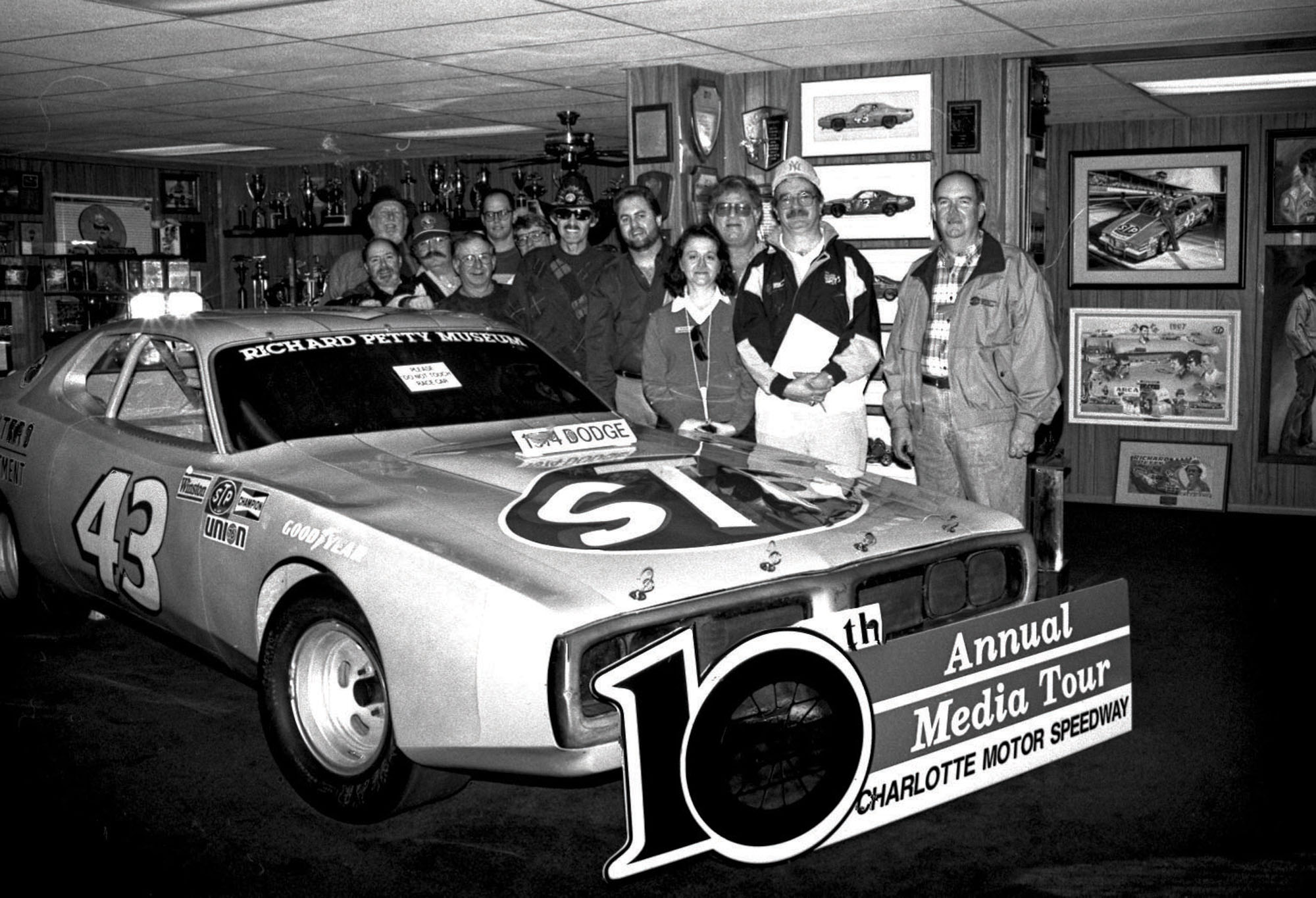 Old photo of Deb Williams and a group posing in front of a car with a sign that reads “10th Annual Media Tour, Charlotte Motor Speedway."