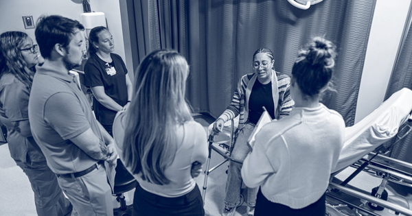 A photo of health sciences students standing around a hospital bed and listening to a standardized patient. 