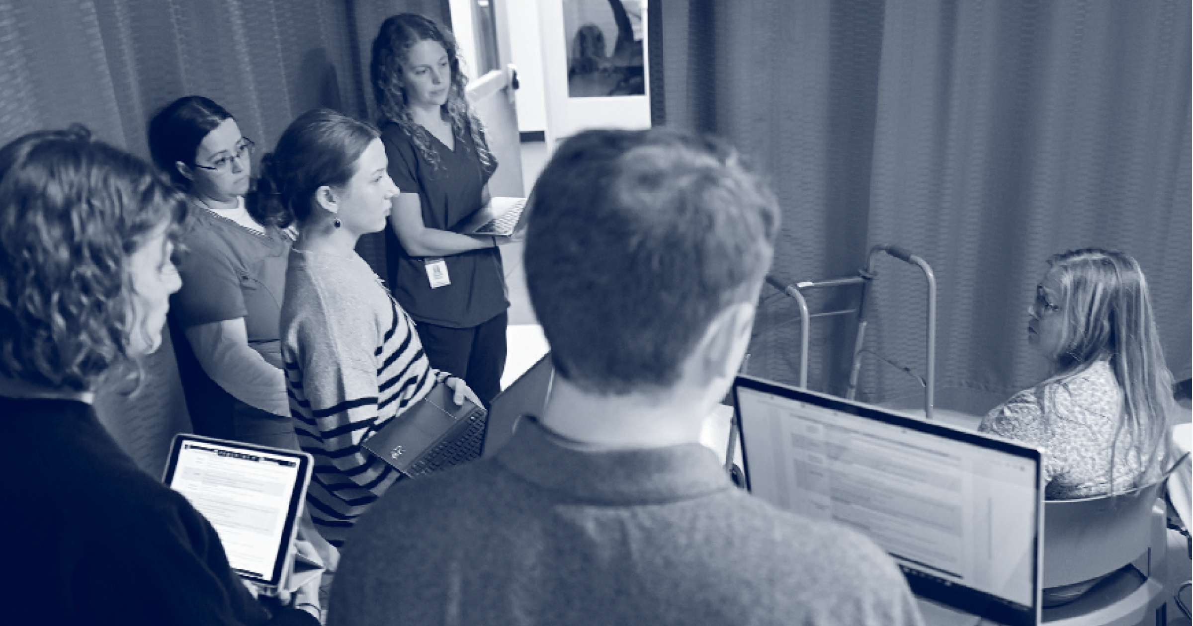 A photo of a standardized patient sitting on a hospital bed and talking to health sciences students.