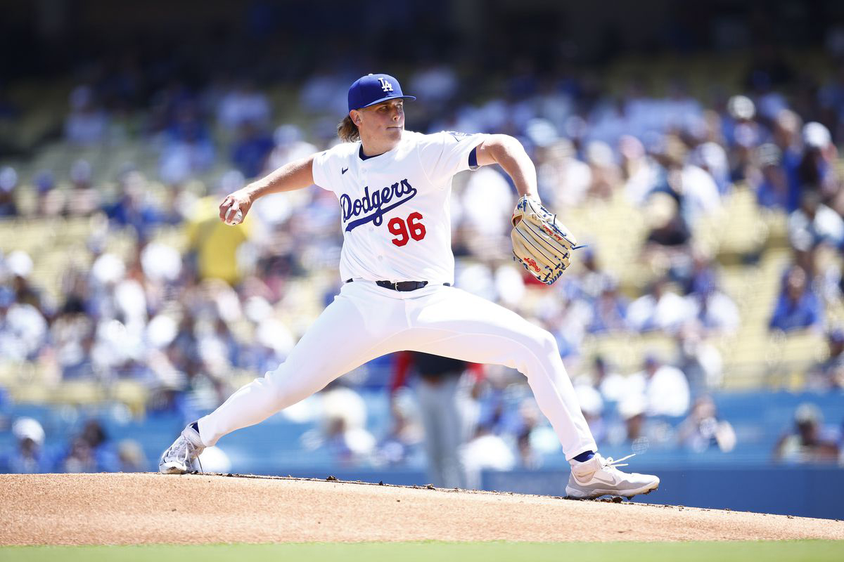 A baseball pitcher in a white Los Angeles Dodgers uniform throwing a pitch from the mound, with fans visible in the stadium seats behind him.