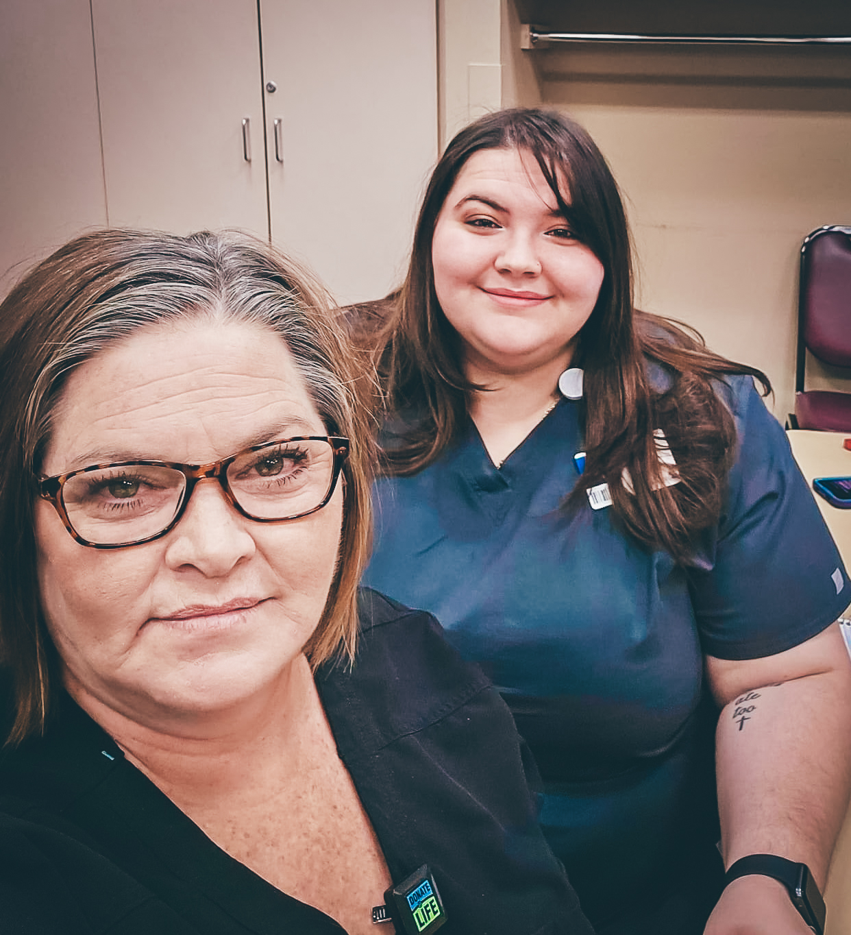 Two health care professionals wearing scrubs, one in glasses, taking a selfie in a clinical or classroom setting.