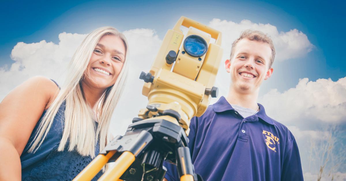 Two students stand outdoors behind a yellow surveying instrument, both smiling against a backdrop of a bright blue sky.