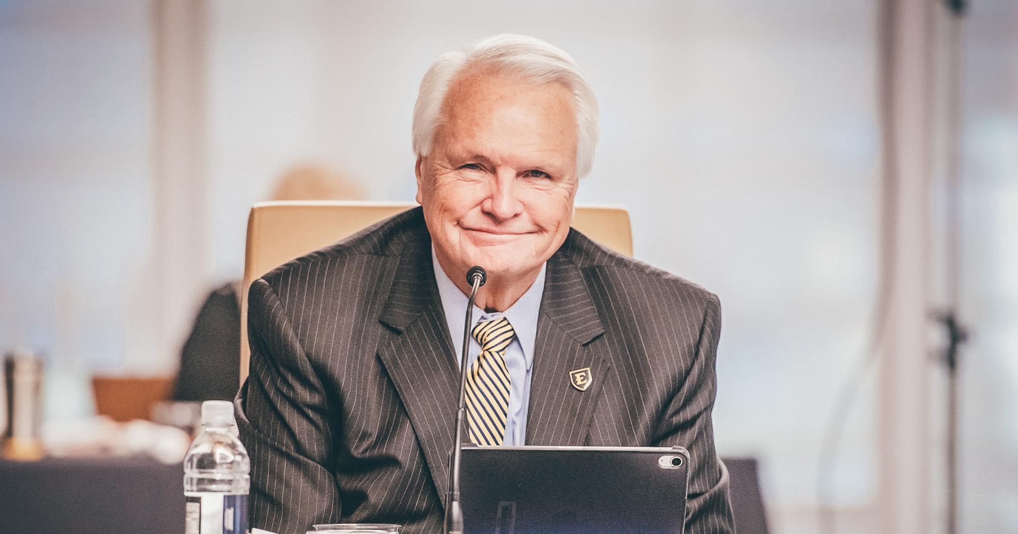 A man in a suit sits at a conference table with a microphone and nameplate, smiling confidently.