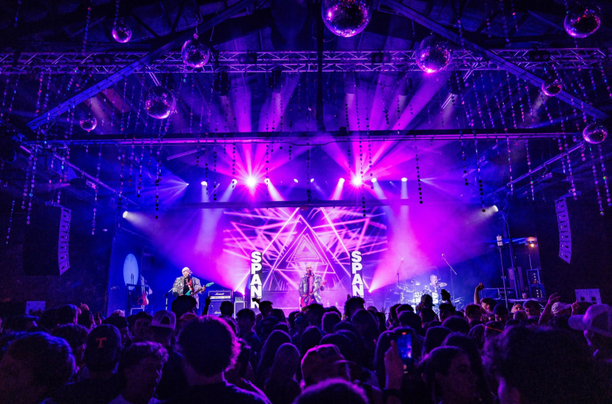 Live concert view of band Spank performing on a stage illuminated by vibrant purple lighting and disco balls, facing a crowded audience.