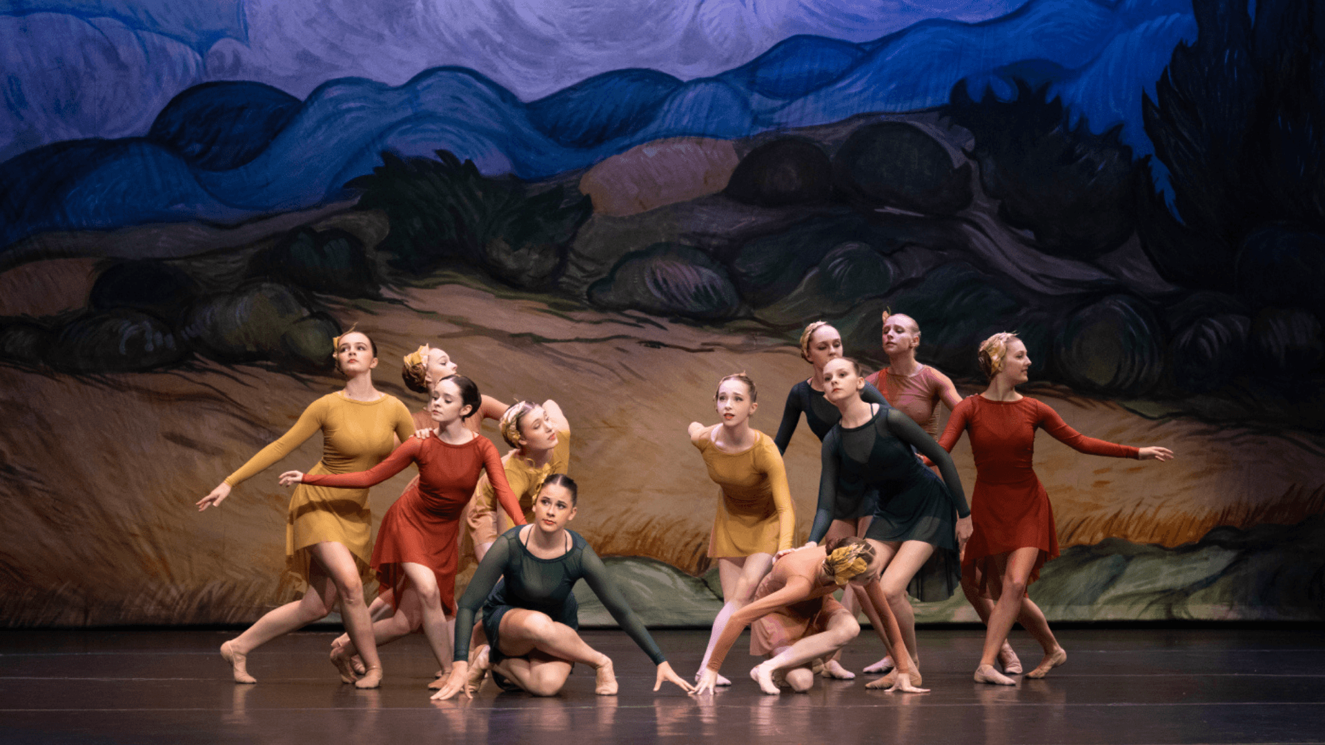 Eleven female dancers in variations of mustard yellow, burnt orange, and emerald green long sleeve short length dresses. They are performing on a stage with a handpainted background of rolling hay fields and mountains. All of the dancers have serious looks on their faces with some standig with their legs crossed, and some crouched towards the stage floor.
