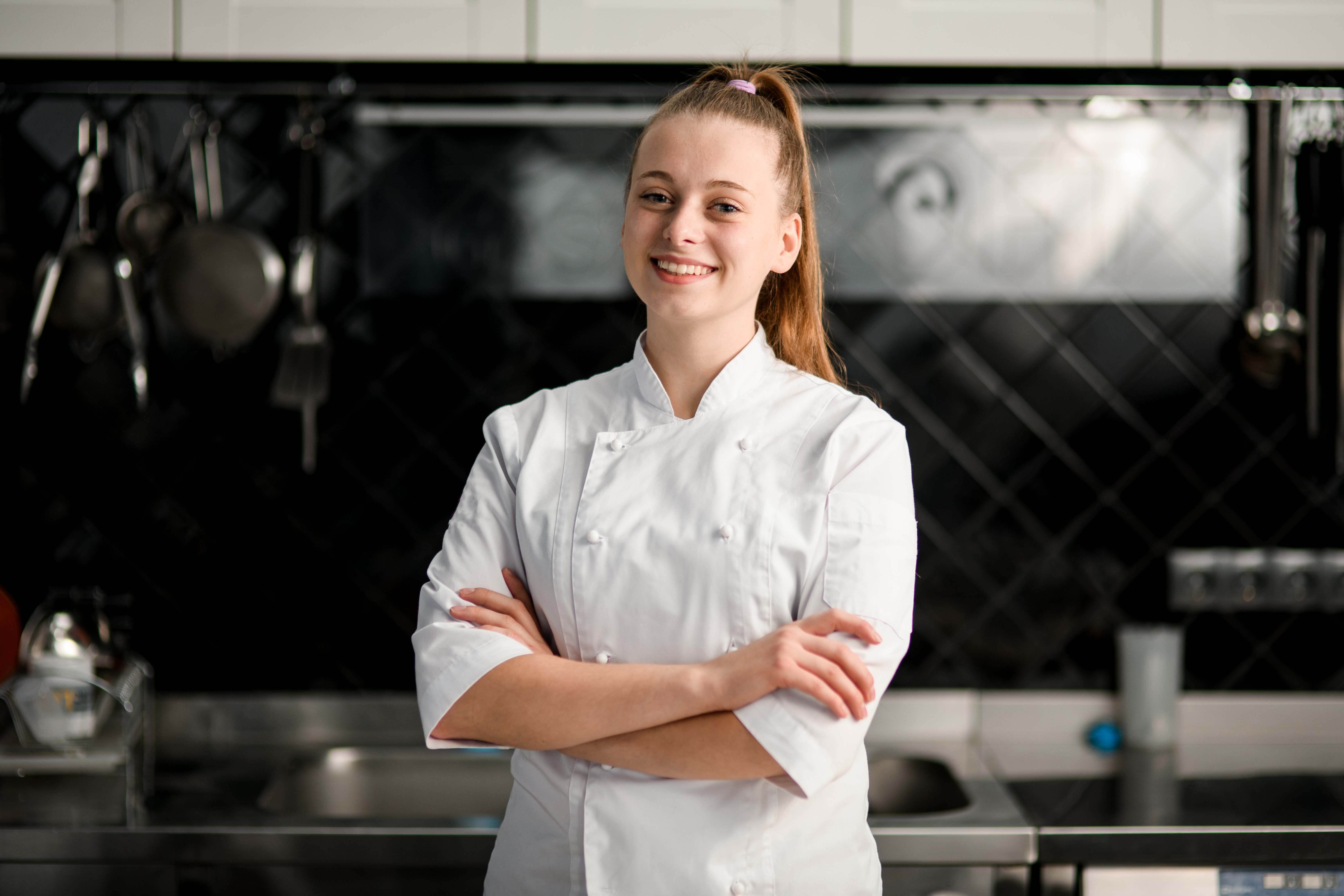 A woman smiles and crosses her arms in an industrial kitchen. She is wearing a white smock. 