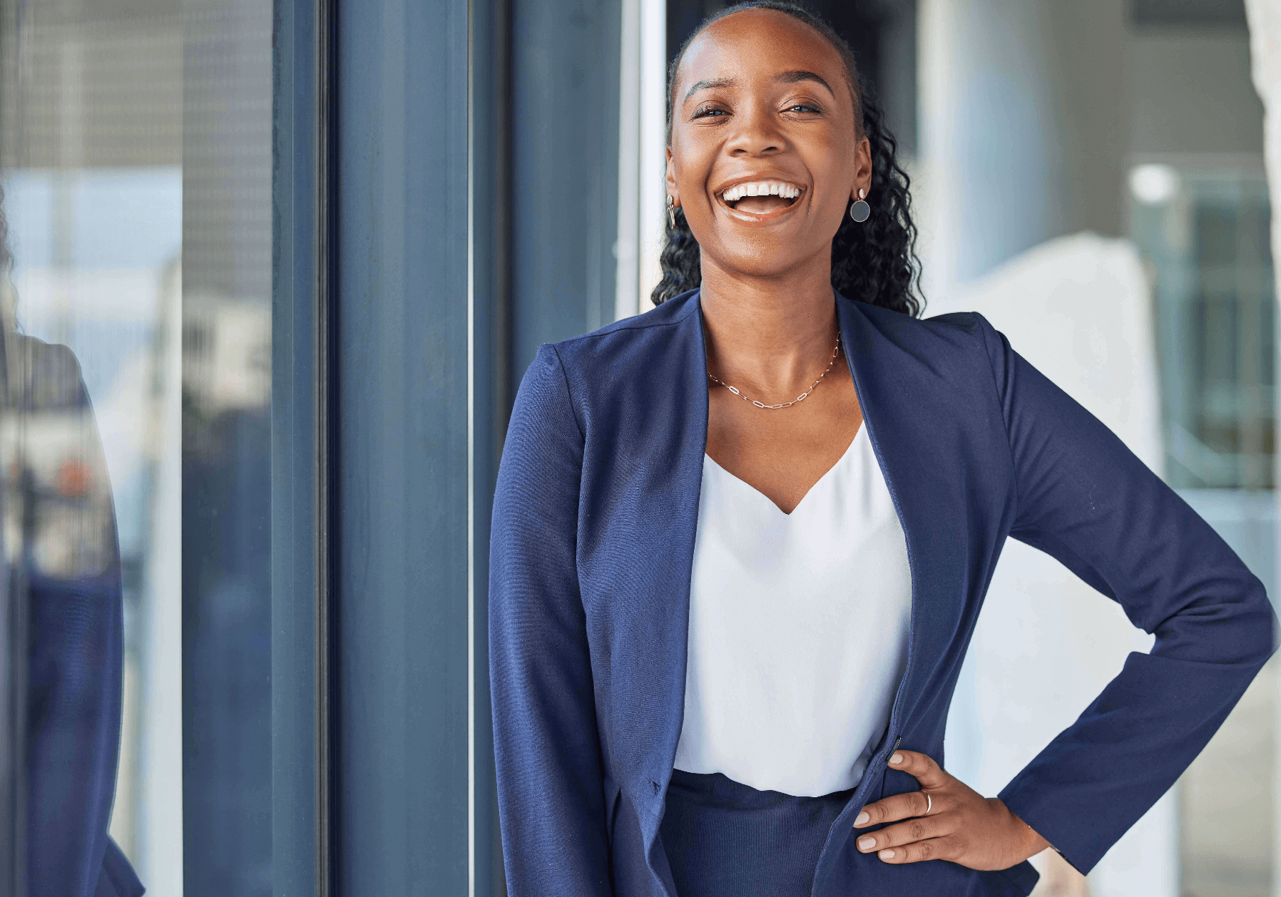 A woman poses for the camera. She is in an office setting and wearing a tailored grey skirt suit. 