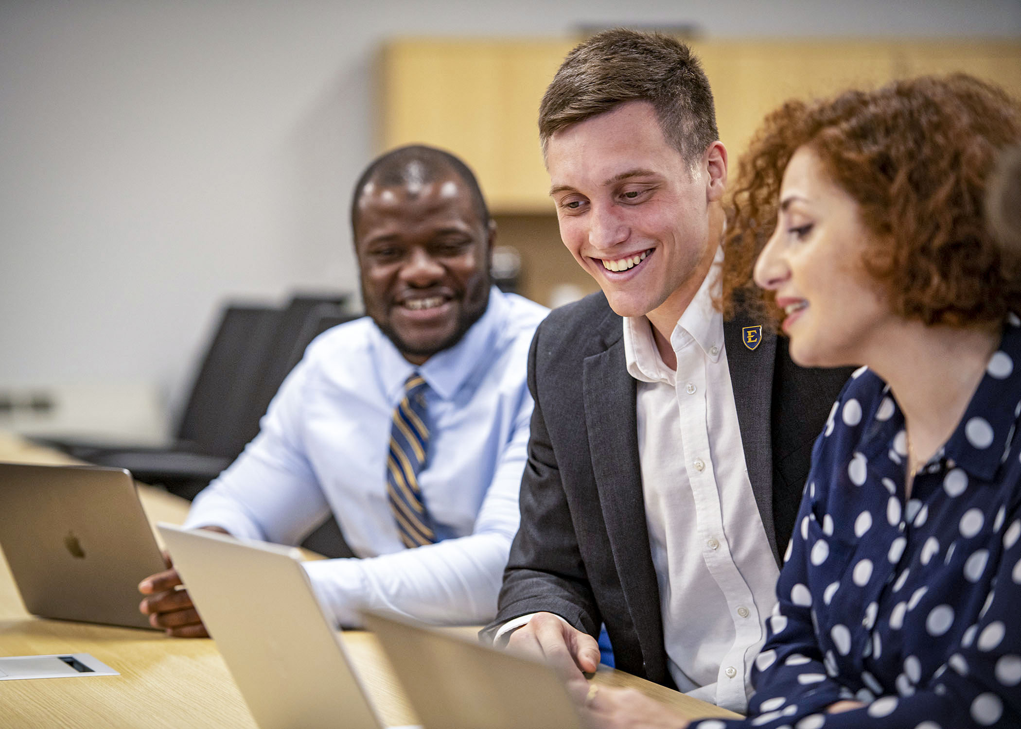 Three people are gathered around a laptop in a classroom.