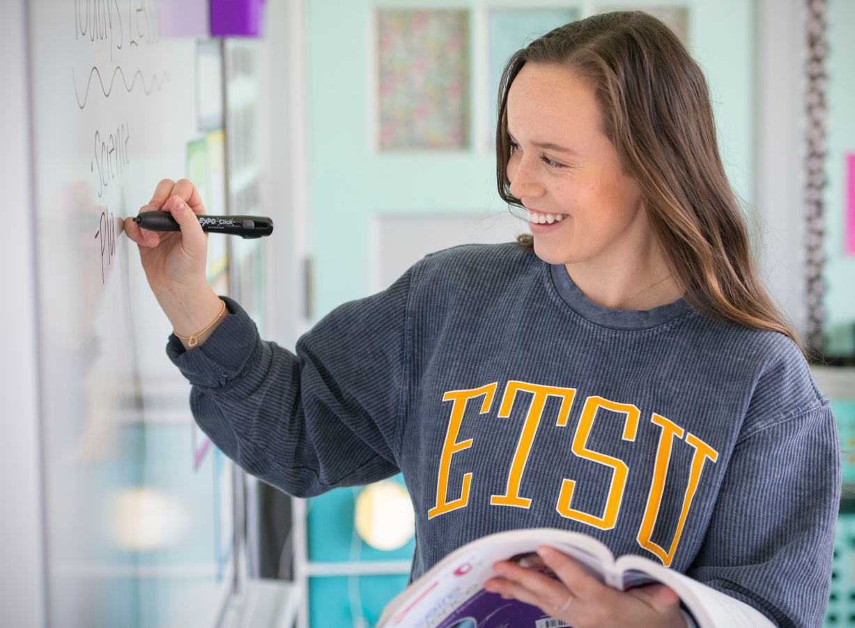 A young woman stands at a white board. She is writing and holding a textbook. 