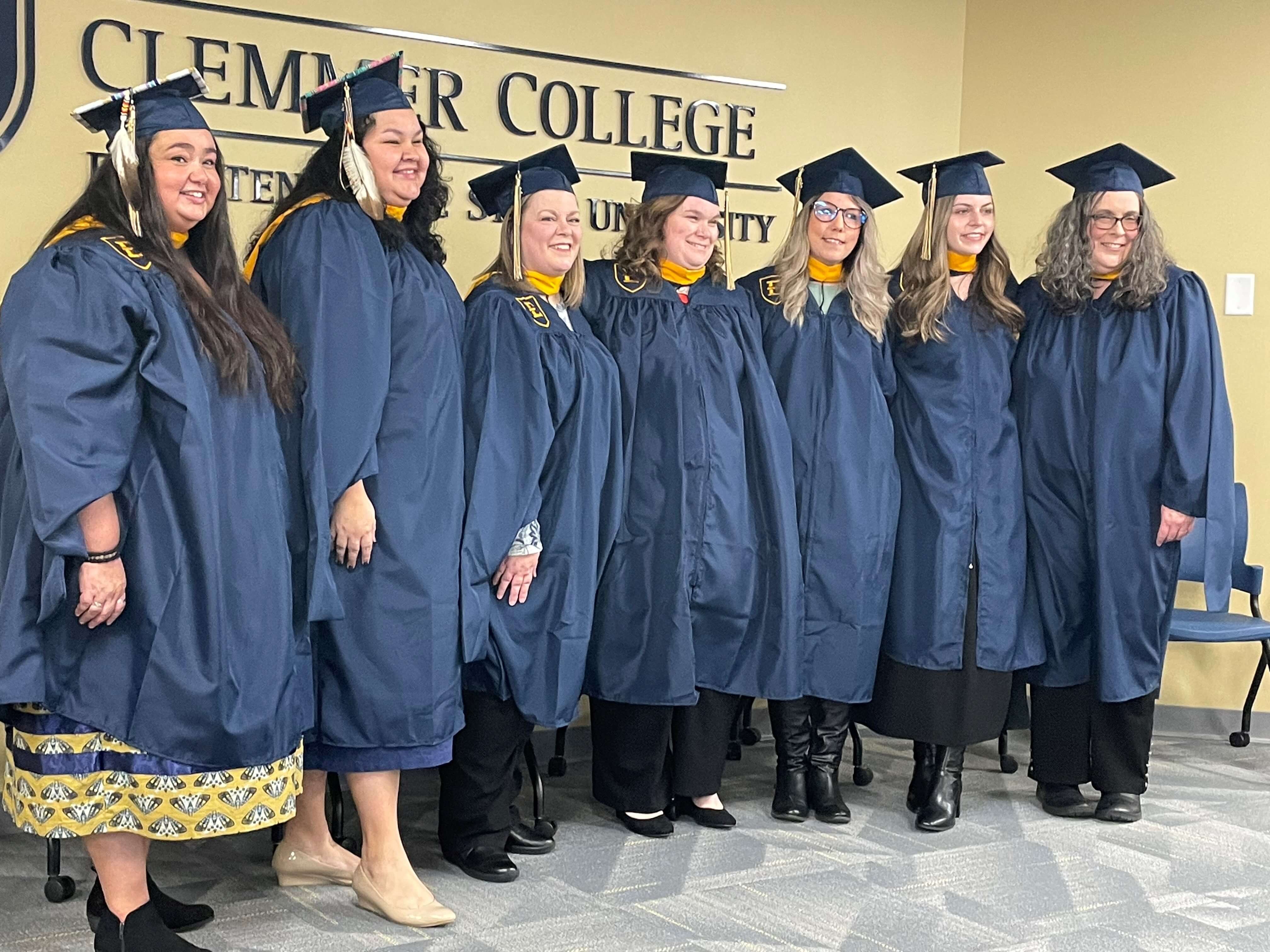 A group of seven graduates pose for a picture. Their arms are around each other and they are smiling.