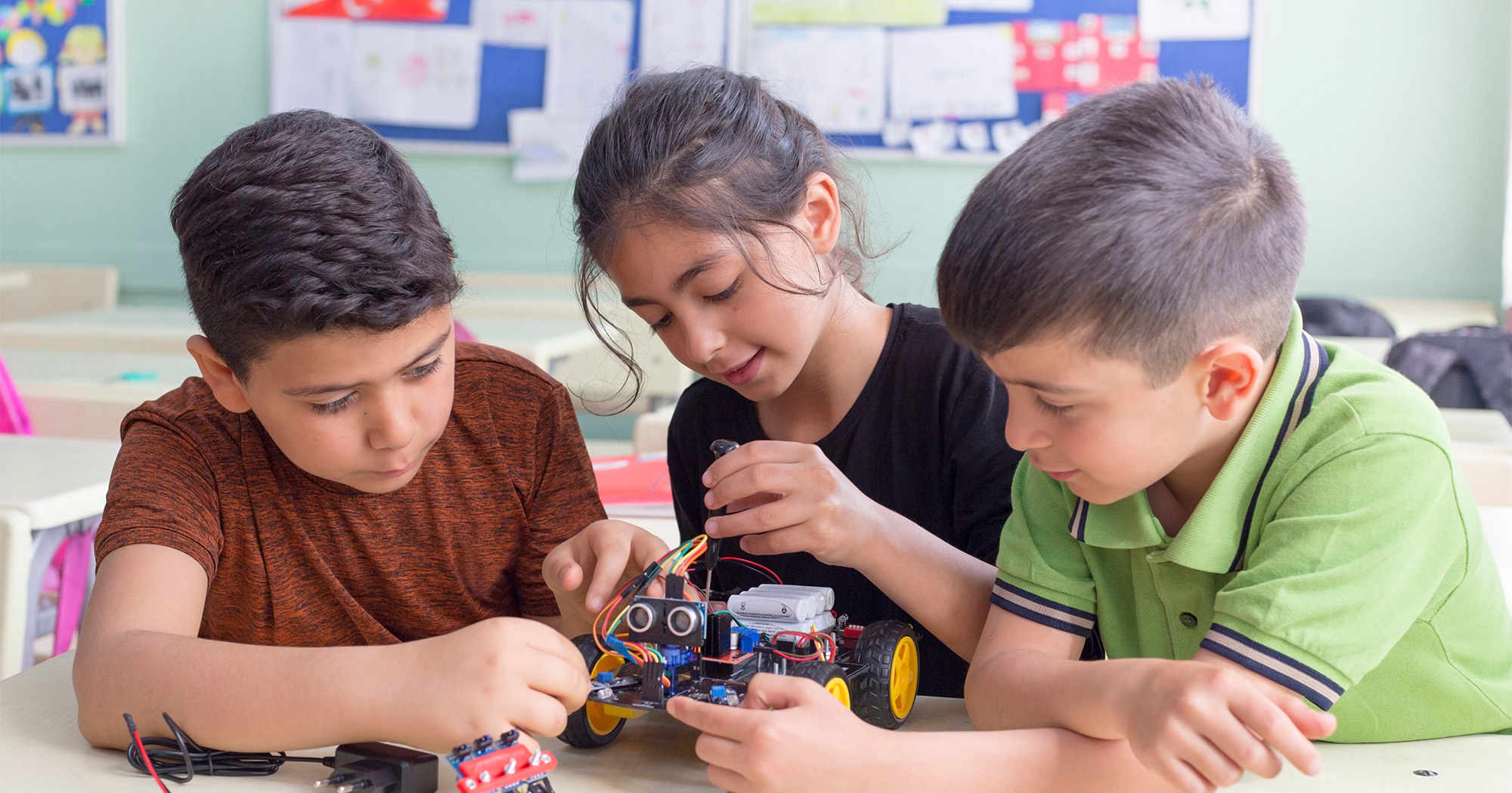 Three 7 to 9 year olds sit in a classroom at a desk. They are working together on a small robotic machine. 