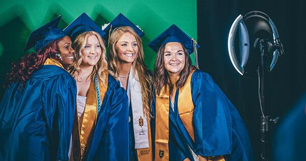 Four women stand in front of a selfie station. They are dressed in gradaution robes.